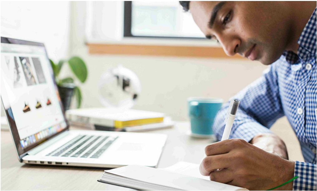 Image of man writing on note pad while a laptop is open